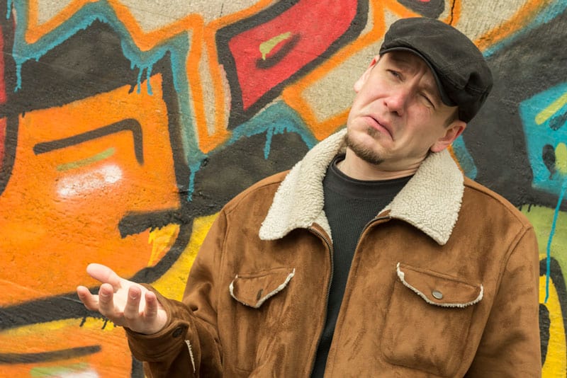 Man in a leather jacket and cap with a quizzical expression, holding a hand up looking like he's trying to decide between options.